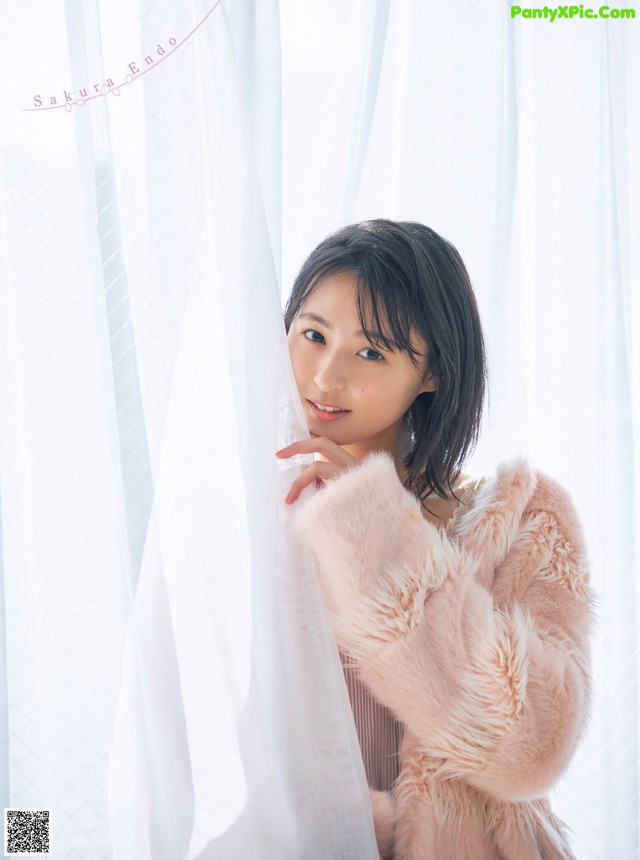 A woman in a pink fur coat leaning against a curtain.